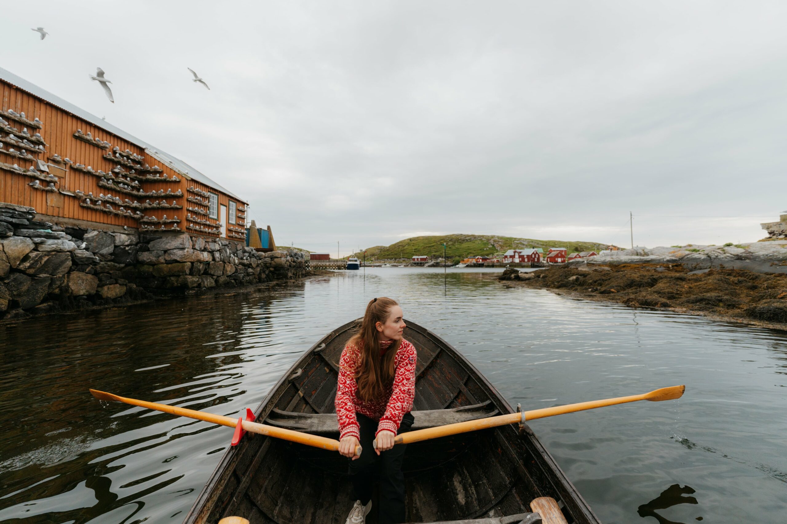 Jente med rød ullgenser sitter i robåt med årene opp av vannet. Bak henne ser vi historiske bygnigner i fiskevær og måser, samt litt svaberg.